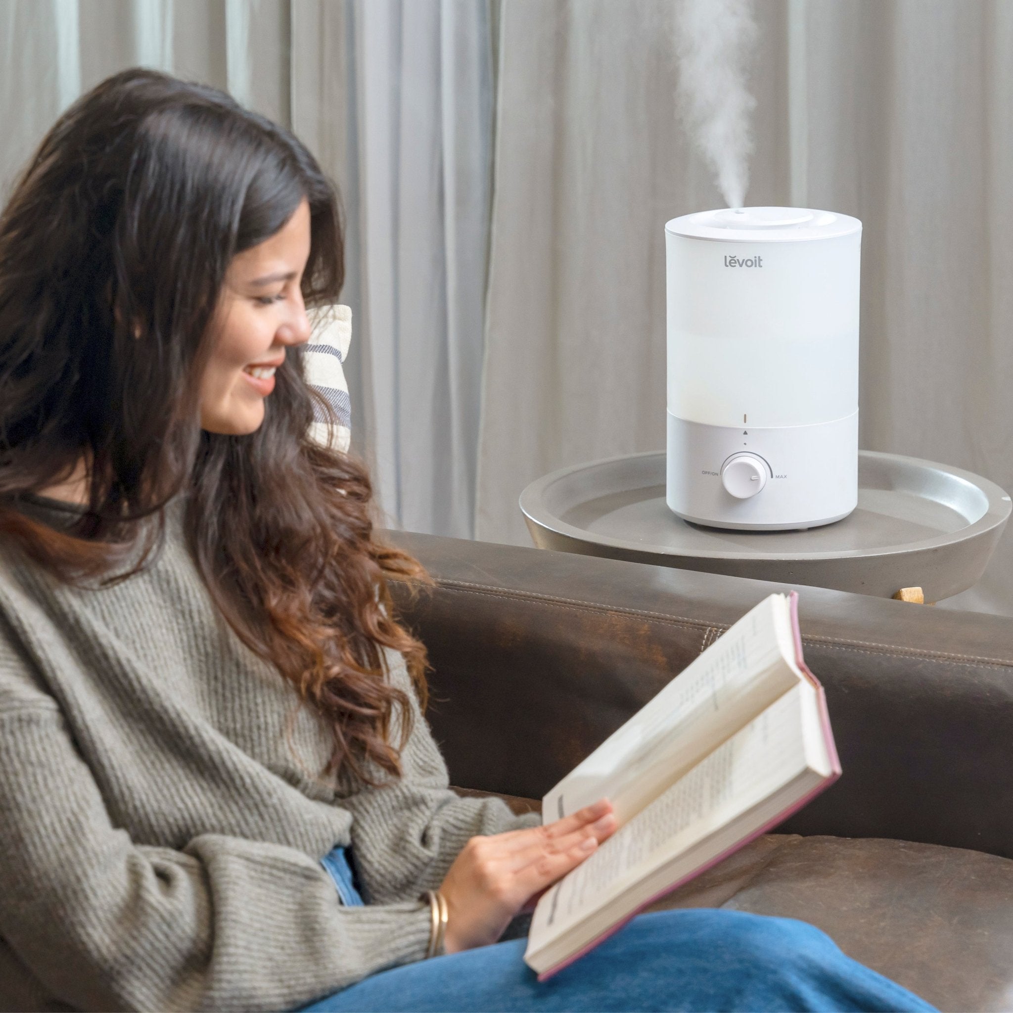 A woman relaxing and reading a book next to the Levoit Dual 150 Ultrasonic Cool Mist Humidifier, with mist gently rising from the unit.